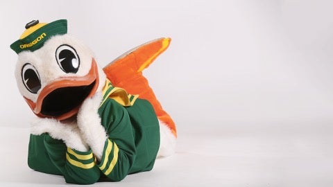 UO Duck mascot laying on the ground, head in hands, looking at the camera