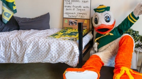 UO Duck mascot sitting on a chair with feet on a table, in a dormitory room setting